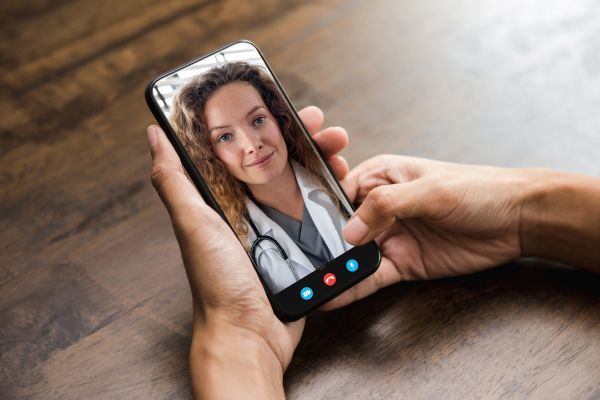 A person holding a smartphone showing a female doctor on a video call, illustrating telehealth and remote healthcare services.